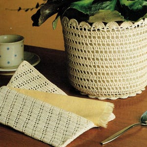 May include: A cream-colored crocheted plant pot cover with a scalloped top edge. A matching crocheted coaster and two crocheted placemats are on a wooden surface. A cup and saucer with blue polka dots are in the background.