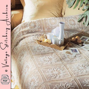 May include: A cream-colored, floral-patterned crochet blanket covers a bed. A wooden tray holds white mugs, a tall white vase, and pastries. The blanket features a repeating square design with flower motifs. The image also includes the text "Vintage Stitchery Archives".