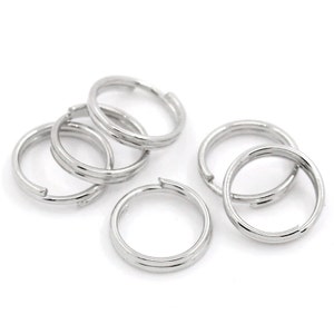 May include: Six silver jump rings, also known as key rings, are scattered on a white surface. The rings are all the same size and have a smooth, polished finish.