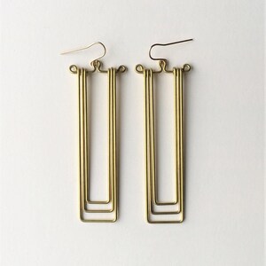 May include: A pair of gold-colored, geometric earrings. Each earring features a rectangular design with three nested rectangles. The earrings have a simple hook closure and are displayed against a white background.