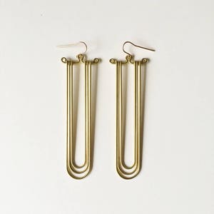 May include: A pair of long, gold-toned earrings with a simple, geometric design. Each earring features three stacked, curved lines that form a U-shape.