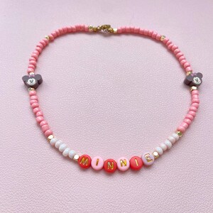 May include: Pink beaded necklace with the name "MINNIE" spelled out in red and pink letter beads. The necklace features two Minnie Mouse head charms and gold-colored accents. A charming accessory.