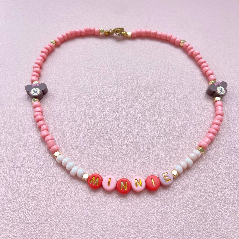 May include: Pink beaded necklace with the name "MINNIE" spelled out in red and pink letter beads. The necklace features two Minnie Mouse head charms and gold-colored accents. A charming accessory.
