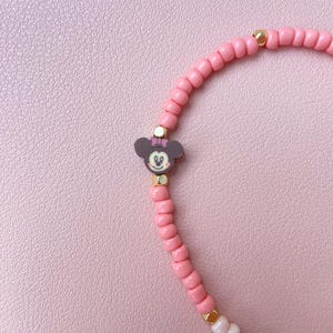 May include: A pink beaded bracelet featuring a Minnie Mouse charm. The bracelet has small, round pink beads with gold-colored accents. The Minnie Mouse charm is dark gray with a pink bow and gold details. The bracelet is on a pink background.
