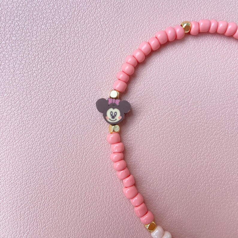 May include: A pink beaded bracelet featuring a Minnie Mouse charm. The bracelet has small, round pink beads with gold-colored accents. The Minnie Mouse charm is dark gray with a pink bow and gold details. The bracelet is on a pink background.