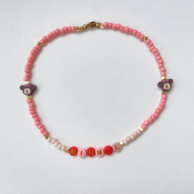 May include: Pink beaded necklace with the name "MINNIE" spelled out in colorful letter beads. The necklace features two Minnie Mouse head charms and gold-colored accents. A charming accessory.