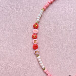 May include: A pink beaded bracelet with the name "WINNIE" spelled out in red and pink letter beads with gold accents. The bracelet features a mix of pink, white, and gold beads.