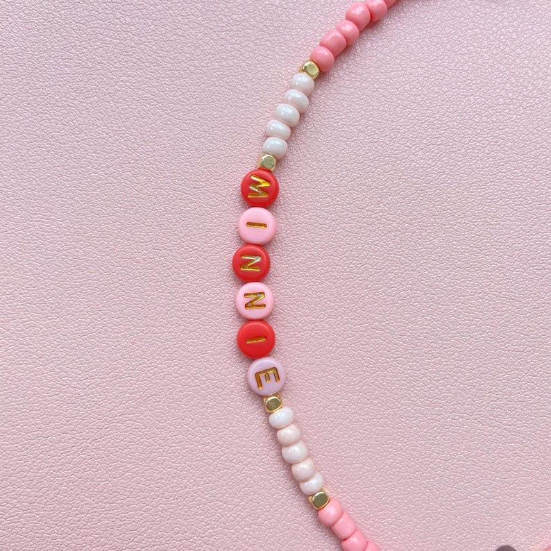 May include: A pink beaded bracelet with the name "WINNIE" spelled out in red and pink letter beads with gold accents. The bracelet features a mix of pink, white, and gold beads.