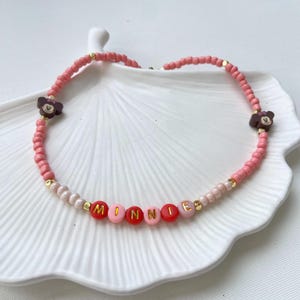 May include: A pink beaded bracelet with the name "MINNIE" spelled out in red, pink, and gold letter beads. The bracelet features two brown Minnie Mouse head charms and gold accent beads.