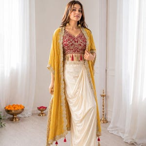 May include: A three-piece ensemble featuring a maroon sleeveless crop top with intricate gold embroidery and small red tassels, a cream-coloured draped skirt, and a long mustard yellow open-front duster jacket with gold lace trim and red tassels.