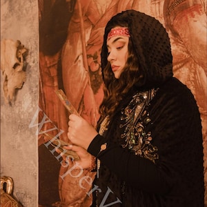 Puede incluir: Capa negra con capucha con intrincados bordados dorados y de joyas. La capa presenta una textura punteada y se usa con una diadema roja. El texto "WhisperVeil" es visible. El fondo es una pintura de estilo renacentista.