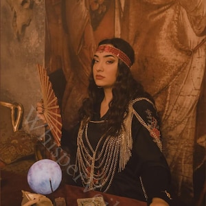 Peut inclure: Une femme v&ecirc;tue d'une robe noire avec des cha&icirc;nes dor&eacute;es et un bandeau rouge. Elle tient un &eacute;ventail rouge et est assise &agrave; une table avec un globe lumineux, des cartes de tarot et d'autres objets mystiques. L'arri&egrave;re-plan est un mur s&eacute;pia.