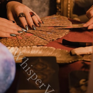 Puede incluir: Primer plano de cartas del tarot abanicadas sobre una superficie roja. La imagen presenta manos con anillos, sosteniendo y organizando las cartas. El texto "Whisper Veil" es visible en la esquina inferior izquierda. El fondo est&aacute; borroso, con un cristal y estrellas.