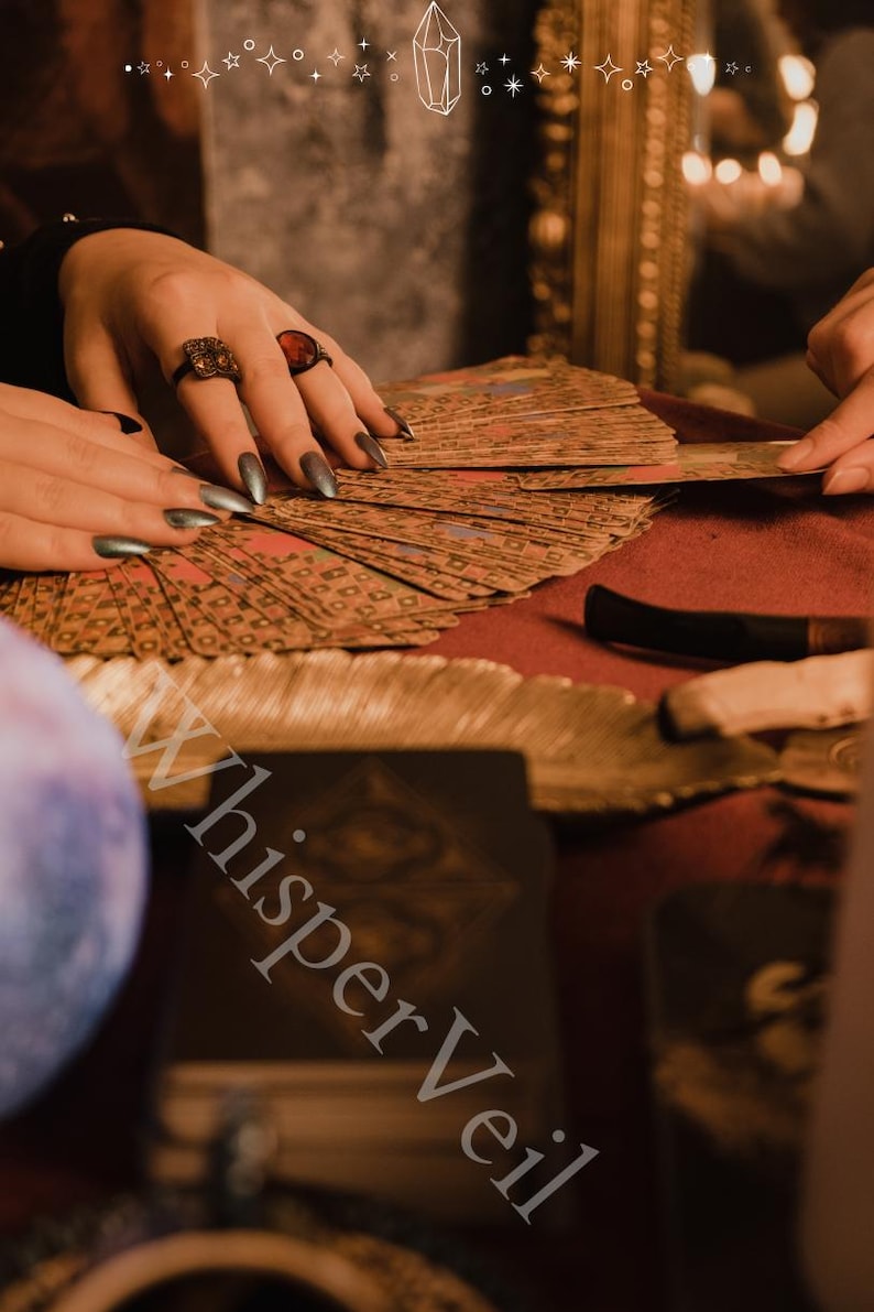 Puede incluir: Primer plano de cartas del tarot abanicadas sobre una superficie roja. La imagen presenta manos con anillos, sosteniendo y organizando las cartas. El texto "Whisper Veil" es visible en la esquina inferior izquierda. El fondo est&aacute; borroso, con un cristal y estrellas.