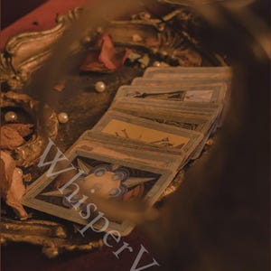 May include: A stack of tarot cards with ornate illustrations, arranged on a red surface. The cards are surrounded by pearls, dried rose petals, and a decorative gold-colored tray. The text "Whisper Veil" is visible.