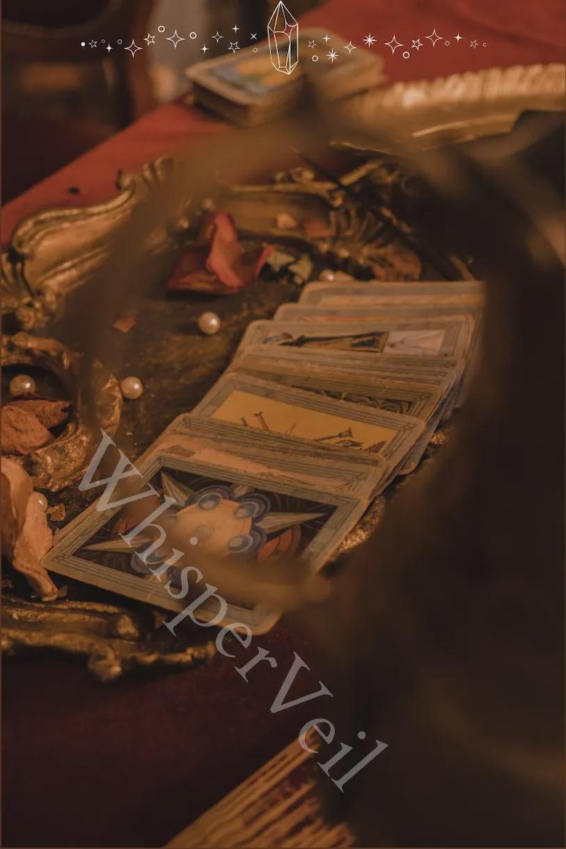 May include: A stack of tarot cards with ornate illustrations, arranged on a red surface. The cards are surrounded by pearls, dried rose petals, and a decorative gold-colored tray. The text "Whisper Veil" is visible.