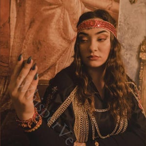 Puede incluir: Una mujer con una diadema roja con lentejuelas y una blusa negra adornada con cadenas doradas. La imagen tiene una est&eacute;tica vintage, con una paleta de colores c&aacute;lidos y un fondo decorativo. La mujer tambi&eacute;n lleva una pulsera roja.