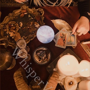 Puede incluir: Una escena m&iacute;stica con una luz en forma de luna brillante, cartas del tarot, velas y una bandeja decorativa. La imagen presenta una superficie roja oscura con varios objetos asociados con la adivinaci&oacute;n y la espiritualidad. La palabra "Whisper Veil" es visible.