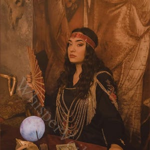 May include: A woman in a black dress with gold chain accents and a red headband. She holds a red fan and sits at a table with a glowing orb, tarot cards, and other mystical items. The image has a vintage, mystical theme.