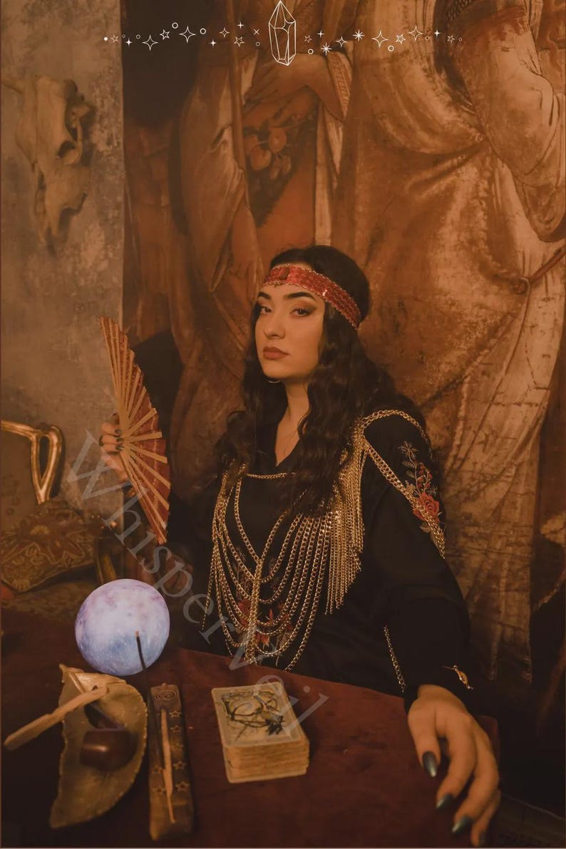 May include: A woman in a black dress with gold chain accents and a red headband. She holds a red fan and sits at a table with a glowing orb, tarot cards, and other mystical items. The image has a vintage, mystical theme.