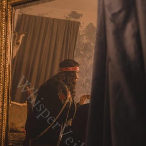 May include: A person in a black embroidered garment with gold chain accents, seated in front of a mirror. The person wears a red headband. The image has the text "Whisper Veil" overlaid on it.