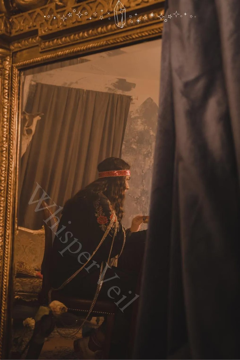 May include: A person in a black embroidered garment with gold chain accents, seated in front of a mirror. The person wears a red headband. The image has the text "Whisper Veil" overlaid on it.