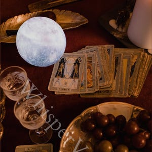 Puede incluir: Una escena m&iacute;stica con una luna brillante, cartas del tarot y una vela encendida sobre una superficie roja. Tambi&eacute;n hay dos copas de vino y un cuenco de uvas. La imagen evoca la adivinaci&oacute;n y las pr&aacute;cticas espirituales.