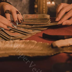 May include: A close-up of a tarot card reading scene. Hands with long, silver nails are arranging a deck of cards on a red surface. The cards are vintage-looking, with detailed illustrations. A feather and a knife are also visible.