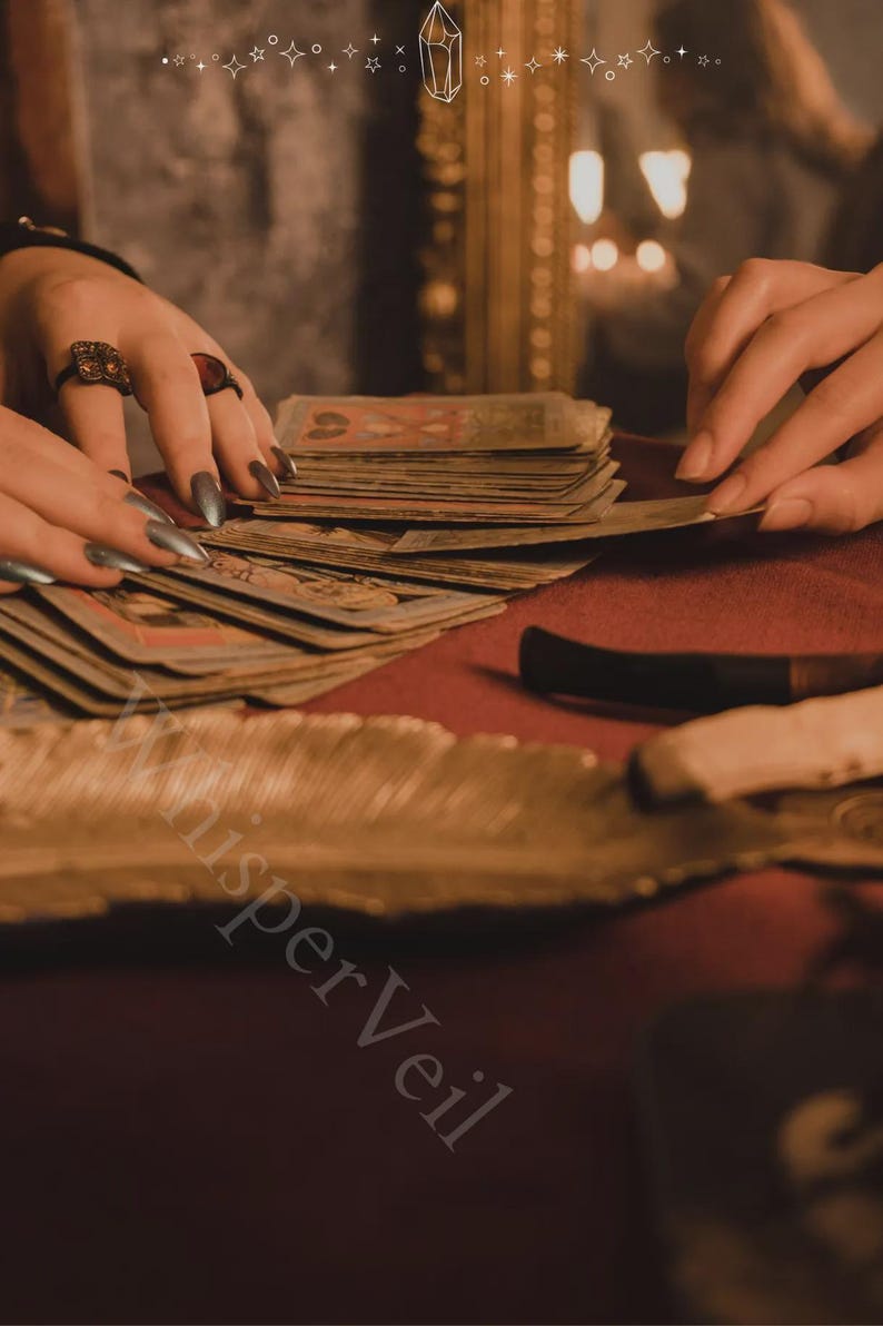 May include: A close-up of a tarot card reading scene. Hands with long, silver nails are arranging a deck of cards on a red surface. The cards are vintage-looking, with detailed illustrations. A feather and a knife are also visible.