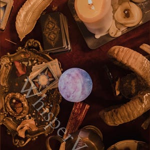 Puede incluir: Un bodeg&oacute;n m&iacute;stico con cartas del tarot, velas encendidas y una pluma decorativa. Un orbe p&uacute;rpura brillante y objetos de metal antiguos est&aacute;n dispuestos sobre una superficie rojo oscuro, creando una est&eacute;tica vintage.