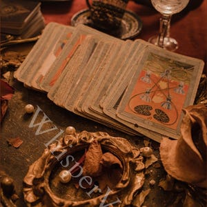 Puede incluir: Una baraja de cartas de tarot vintage con un dise&ntilde;o naranja y azul, abanicada sobre una mesa. Las cartas est&aacute;n desgastadas y envejecidas, con un marco dorado ornamentado y perlas. La palabra "WhisperVeil" es visible en la imagen.