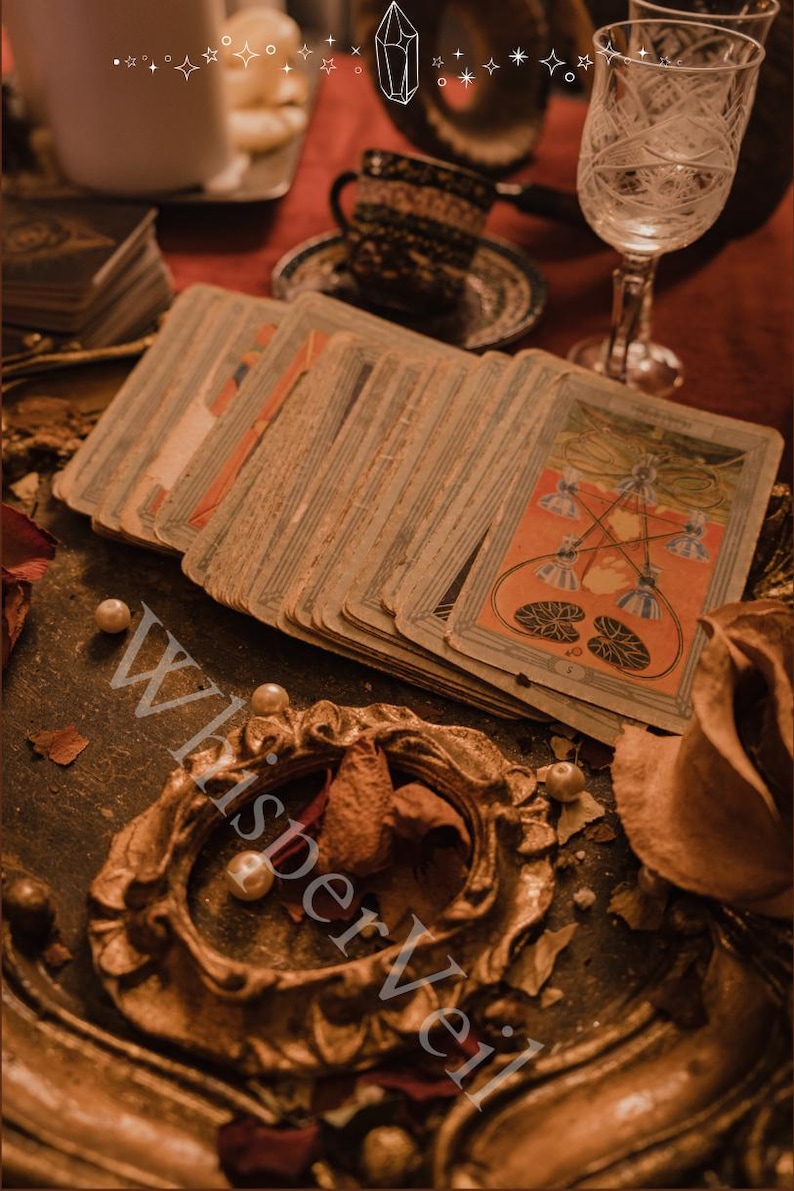 Puede incluir: Una baraja de cartas de tarot vintage con un dise&ntilde;o naranja y azul, abanicada sobre una mesa. Las cartas est&aacute;n desgastadas y envejecidas, con un marco dorado ornamentado y perlas. La palabra "WhisperVeil" es visible en la imagen.