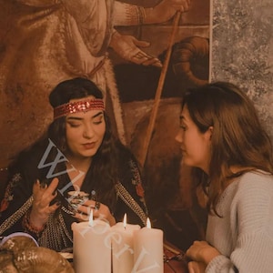 Puede incluir: Dos personas sentadas en una mesa con velas y una calavera de carnero. Una persona lleva una diadema roja y una blusa negra con ribetes dorados. La otra persona lleva un su&eacute;ter gris. La letra "W" es visible.