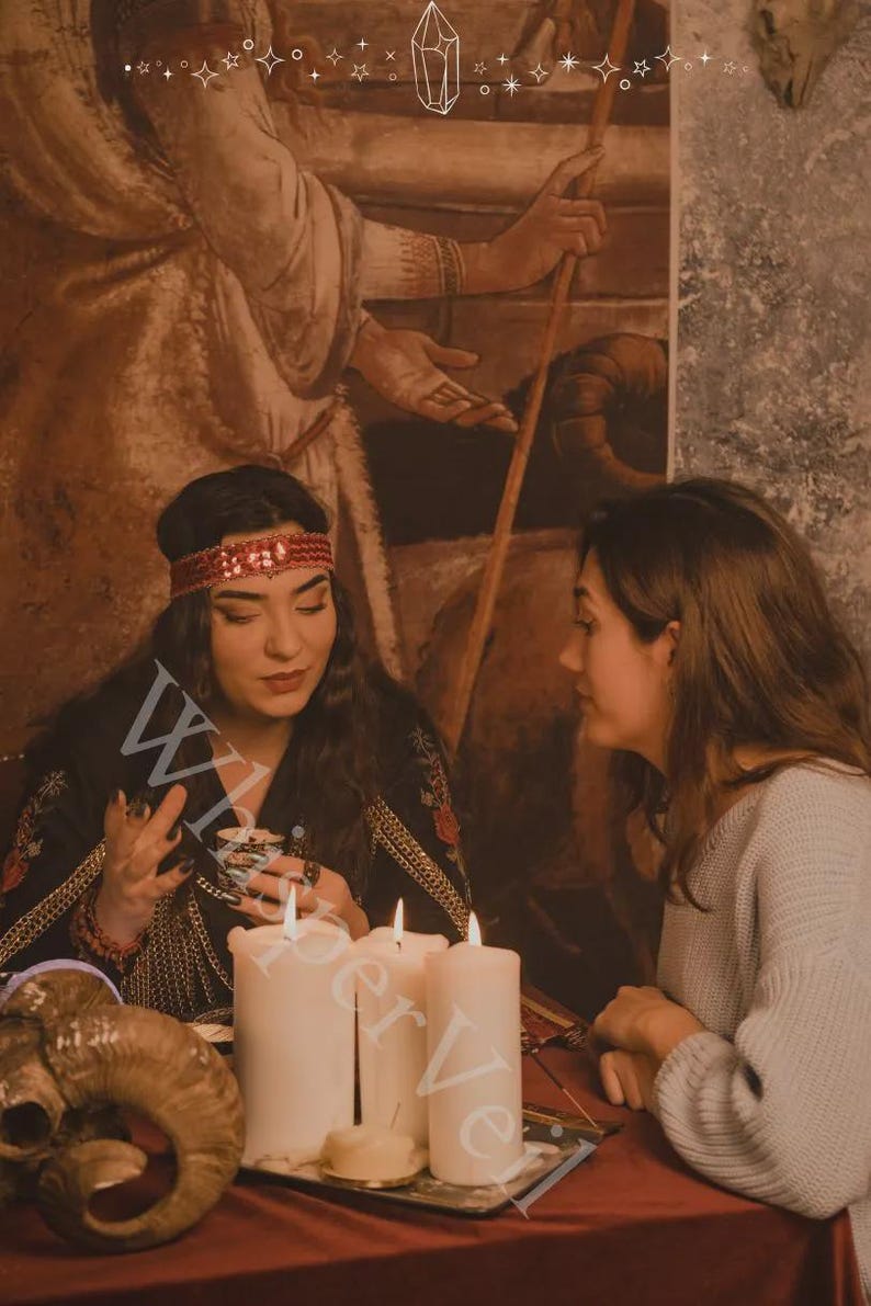 Puede incluir: Dos personas sentadas en una mesa con velas y una calavera de carnero. Una persona lleva una diadema roja y una blusa negra con ribetes dorados. La otra persona lleva un su&eacute;ter gris. La letra "W" es visible.