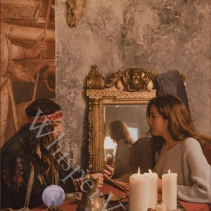 Puede incluir: Imagen atmosf&eacute;rica de dos personas sentadas a una mesa roja, en un entorno m&iacute;stico. La escena incluye velas, un espejo decorativo y un recipiente de lat&oacute;n. La palabra "Whisperveil" es parcialmente visible.