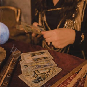 May include: A mystical scene featuring tarot cards laid out on a burgundy surface. The cards have detailed illustrations. A person is holding more cards, and a decorative fan is visible. The setting has a vintage, esoteric aesthetic.