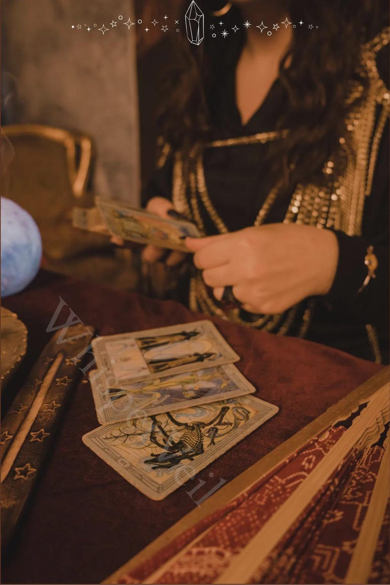 May include: A mystical scene featuring tarot cards laid out on a burgundy surface. The cards have detailed illustrations. A person is holding more cards, and a decorative fan is visible. The setting has a vintage, esoteric aesthetic.