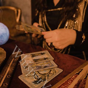 Puede incluir: Cartas del tarot con ilustraciones detalladas dispuestas sobre una superficie de terciopelo burdeos, con una persona sosteniendo m&aacute;s cartas al fondo. Tambi&eacute;n se ven un porta incienso y un abanico decorativo. Tema m&iacute;stico.
