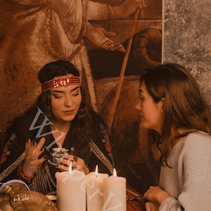 Puede incluir: Una escena de sesi&oacute;n de adivinaci&oacute;n. Dos personas est&aacute;n sentadas a una mesa con velas encendidas, una calavera decorativa y una bandeja. Una persona lleva una diadema roja y una blusa negra bordada. La otra persona lleva un su&eacute;ter gris. El fondo presenta un mural.
