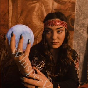 Puede incluir: Una mujer con una blusa negra con detalles rojos y dorados sostiene un orbe azul brillante. Lleva una diadema roja con lentejuelas y collares de cadena dorada. La imagen tiene el texto "WhisperVeil".