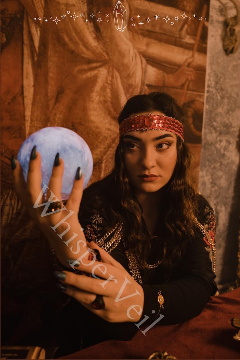 Puede incluir: Una mujer con una blusa negra con detalles rojos y dorados sostiene un orbe azul brillante. Lleva una diadema roja con lentejuelas y collares de cadena dorada. La imagen tiene el texto "WhisperVeil".
