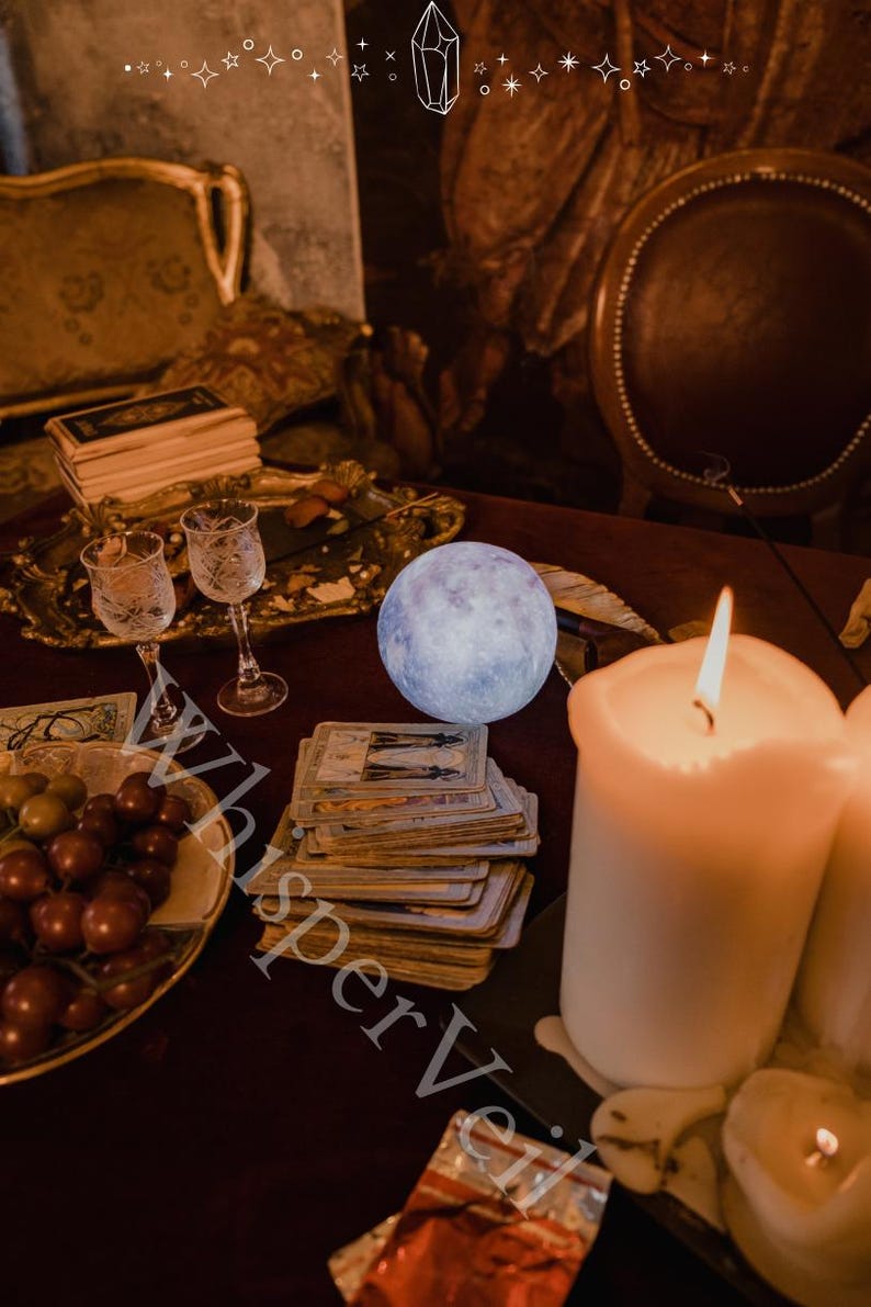 Puede incluir: Una escena m&iacute;stica con un orbe azul brillante, cartas del tarot, velas encendidas y copas de vino. Tambi&eacute;n hay un plato de uvas y una bandeja decorativa. La imagen tiene un ambiente c&aacute;lido y acogedor, con el texto "Whisper Veil" parcialmente visible.