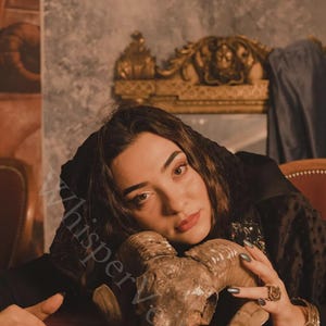 May include: A ram skull with large horns rests on a red surface. The skull is brown and tan, with visible bone texture. The image has a vintage aesthetic, with a dark background and soft lighting. The overall theme is gothic or occult.