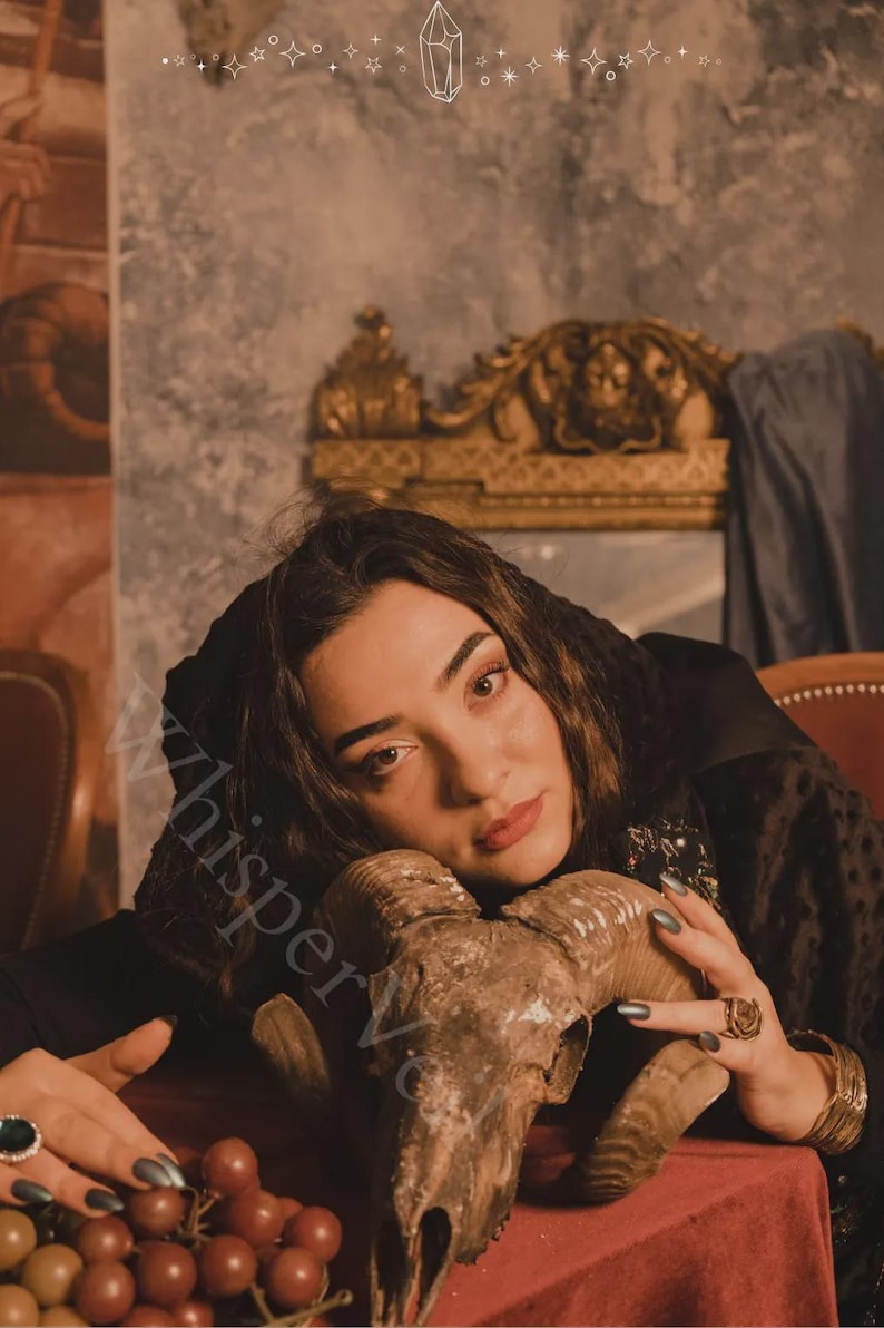 May include: A ram skull with large horns rests on a red surface. The skull is brown and tan, with visible bone texture. The image has a vintage aesthetic, with a dark background and soft lighting. The overall theme is gothic or occult.