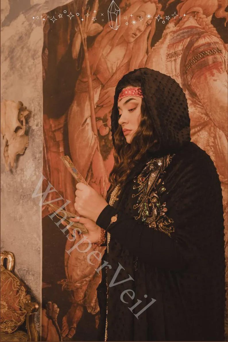May include: A woman wearing a black hooded cloak with ornate gold and jewel embroidery. She is holding a decorative fan. The background features a Renaissance-style painting with warm tones and the text "Whisper Veil".