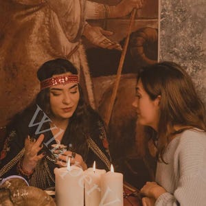 May include: An image of two people in a dimly lit room, with candles and a ram skull on a table. One person is wearing a red headband and a black top with gold trim. The other person is wearing a gray sweater. The word "W" is visible.
