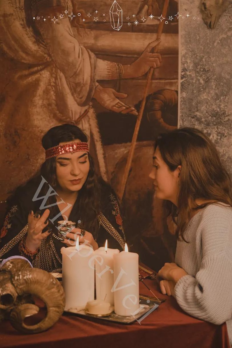 May include: An image of two people in a dimly lit room, with candles and a ram skull on a table. One person is wearing a red headband and a black top with gold trim. The other person is wearing a gray sweater. The word "W" is visible.