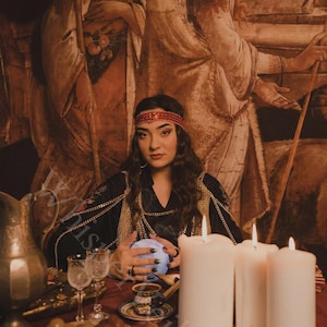 May include: A woman in a black dress with gold chain accents and a red headband sits at a table with a crystal ball. Lit candles, grapes, and glasses are on the table. The background features a vintage painting.