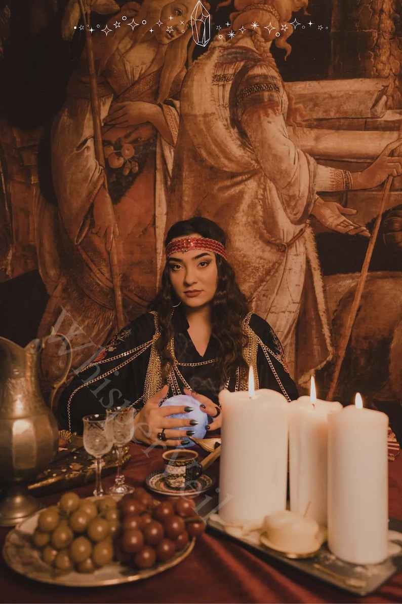 May include: A woman in a black dress with gold chain accents and a red headband sits at a table with a crystal ball. Lit candles, grapes, and glasses are on the table. The background features a vintage painting.