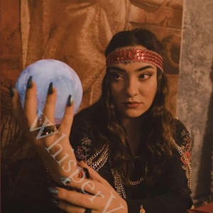 May include: A woman wearing a red sequined headband and a black top with gold chain accents holds a glowing blue orb. The image has a mystical theme, with the text "WhisperVeil" partially visible.